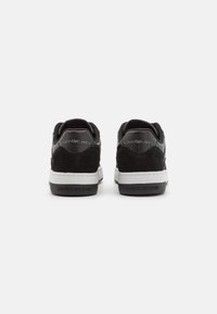 Zapatillas de ante y cuero negras con suelas blancas, que presentan un detalle de marca en la parte trasera y una textura suave. Diseño clásico de corte bajo.