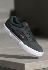 Adidași gri închis cu un upper texturat, logo roz accent, talpă albă plată și șirete negre. Design simplu cu o siluetă low-top.