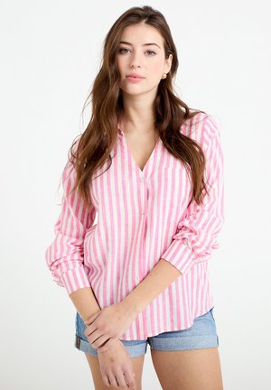 Donna con lunghi capelli castani che indossa orecchini d'oro, una camicia a maniche lunghe a righe verticali rosa e bianche con bottoni e pantaloncini di jeans, in piedi con le braccia incrociate.