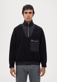 Felpa nera con zip a metà, polsini elasticizzati e un pannello anteriore testurizzato in contrasto. Provvista di tasca con zip e dettaglio del logo.
