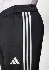 Crne sportske hlače s elastičnim pojasom, koje imaju tri bijele vertikalne pruge i bijeli Adidas logo na donjoj lijevoj strani.
