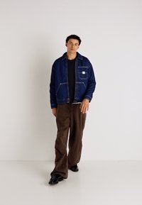 Jeansjack in donkerblauw met een borstzak en contrasterende stiksels, gecombineerd met bruine wijde broek en zwarte schoenen.