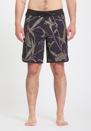Homme portant un short de bain noir avec un imprimé de feuilles tropicales vertes et roses, debout pieds nus devant un fond blanc.