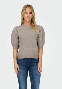 Blonde vrouw in een taupe gebreide trui met pofmouwen en blauwe spijkerbroek, staand tegen een effen lichte achtergrond.