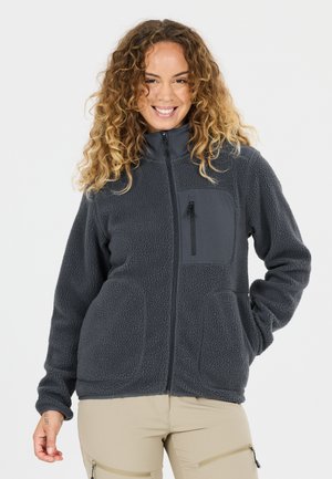 Lächelnde Frau mit lockigem Haar, die eine dunkelgraue Fleecejacke mit Reißverschlusstasche an der Brust und beige Hose mit Reißverschlusstaschen trägt.