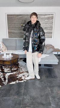 Schwarze, glänzende Daunenjacke mit mehrfarbigen abstrakten Mustern, kombiniert mit cremefarbenen Hosen und weißen Sneakers, stehend auf einem dunklen Fliesenboden.
