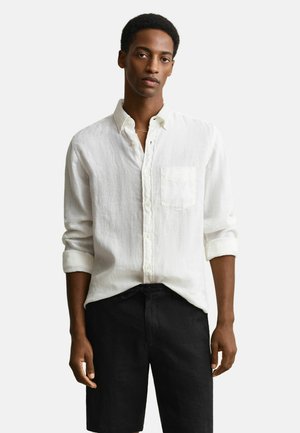 Junger Mann, der ein weißes, langärmeliges Hemd mit Brusttasche und schwarze Shorts trägt, vor einem einfarbigen Hintergrund stehend.