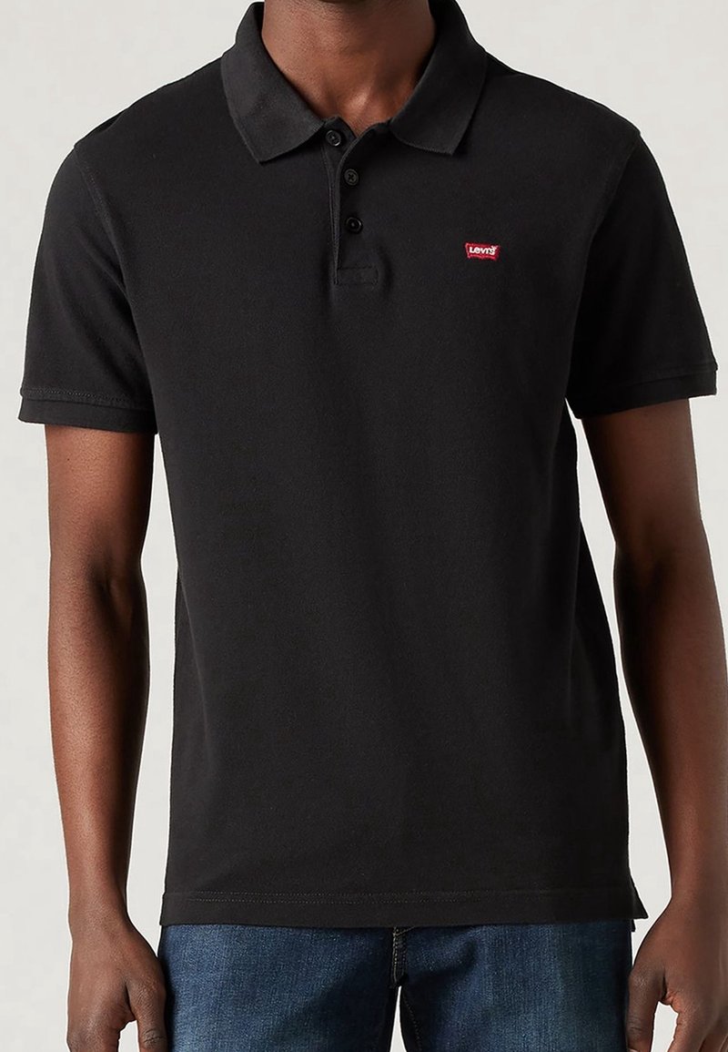 Homme portant un polo noir à manches courtes avec un petit logo rouge Levi's sur la poitrine et un jean bleu sur un fond uni.