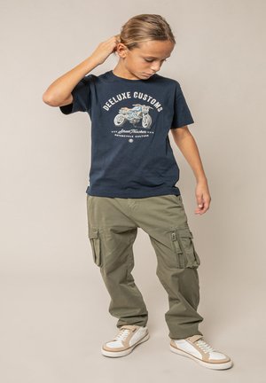 Enfant aux cheveux attachés portant un t-shirt graphique moto bleu marine, un pantalon cargo olive et des baskets blanches, debout devant un fond uni.