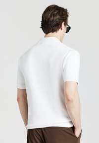 Polo bianco a maniche corte con finitura testurizzata, dotato di colletto classico. Lo sfondo è neutro, esaltando il design della maglietta.