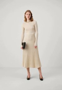 Nais, kes kannab beeži pikka varrukat, ribakudumist ja ääristatud seelikut, hoides musta klambrit ja kandes musti kontsi kuldsete aktsentidega.