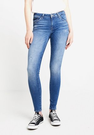 Jeans de mezclilla azul ajustados con cintura de altura media, presentan un ligero desgaste y acentos desgastados, combinados con zapatillas de lona negra.