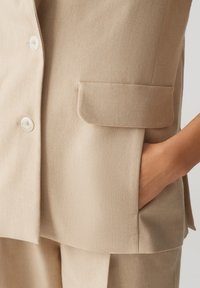 Beige blazer van een textuurstof, met een enkele borstzak, twee witte knopen en een relaxte pasvorm.
