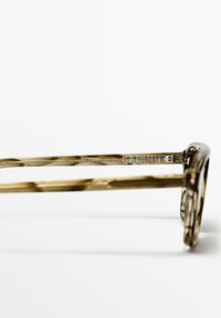 Bruin schildpadkleurig brillenmontuur met een ronde vorm. Getextureerd patroon op de pootjes, voorzien van zilverkleurige hardware en merkgraveerwerk.