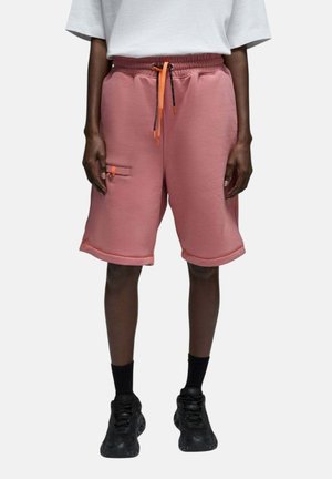 Personne portant un short rose arrivant aux genoux avec un cordon orange, des baskets noires, des chaussettes noires et un t-shirt blanc, debout devant un fond uni.