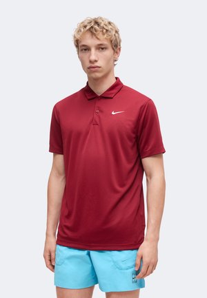 Mladý muž s kučeravými blond vlasmi, oblečený v červené polo Nike a svetlomodré šortky, stojaci pred jednoduchým svetlošedým pozadím.