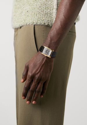 Donkere huidarm met een rechthoekig zwart horloge met een gouden frame en een zwarte leren band, rustend naast kaki broek en een gestructureerde lichte trui.