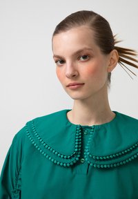 Žalia bliuskutė su per dideliu apykaklės kraštu, puošta tekstūruotais pom-pom akcentais ir mygtukų uždarymu kaklo srityje. Glotni medžiaga, laisvas siluetas.