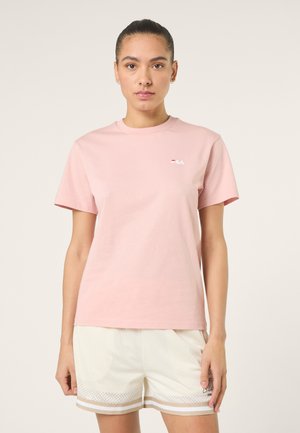 Ung kvinde stående iført en lyserød FILA T-shirt og hvide mesh sportsshorts med beige striber.