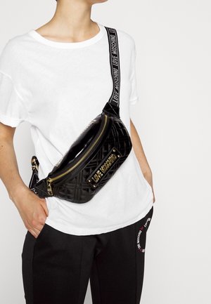Schwarze, gesteppte Crossbody-Tasche mit goldenen Beschlägen, ausgestattet mit einem Reißverschluss und einem Logo-Trageriemen. Wird über einem weißen, lässig geschnittenen Hemd getragen.