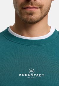 Zbliżenie dolnej części twarzy mężczyzny ubranego w turkusową koszulę na białej podkoszulce, z napisem "KRONSTADT Casual Since 1999" na koszuli.