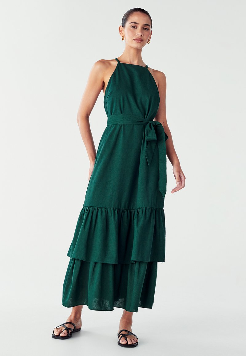 Duga zelena haljina s halter izrezom, oblikovanim gornjim dijelom i slojevitom volanastom suknjom. Ima vezani struk i lagani materijal. Crne sandale.
