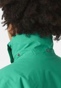 Miętowa wodoodporna kurtka z wysokim kołnierzem, detalami na napy oraz gładką, matową teksturą. Włosy zasłaniają obszar szyi.