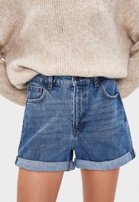 Persona con suéter de punto beige claro metido dentro de pantalones cortos de mezclilla azules de tiro alto y dobladillo vuelto, de pie contra un fondo blanco.