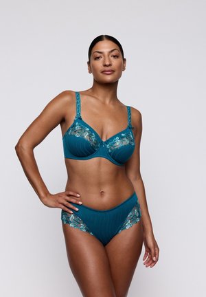 Vrouw met donker haar in tealkleurige kanten lingerie poseert met één hand op haar heup tegen een effen lichte achtergrond.