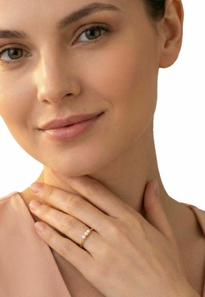 Kobieta z czystą cerą delikatnie dotyka szyi, ma na palcu złoty pierścionek z trzema perłami i nosi jasnoróżowy top.