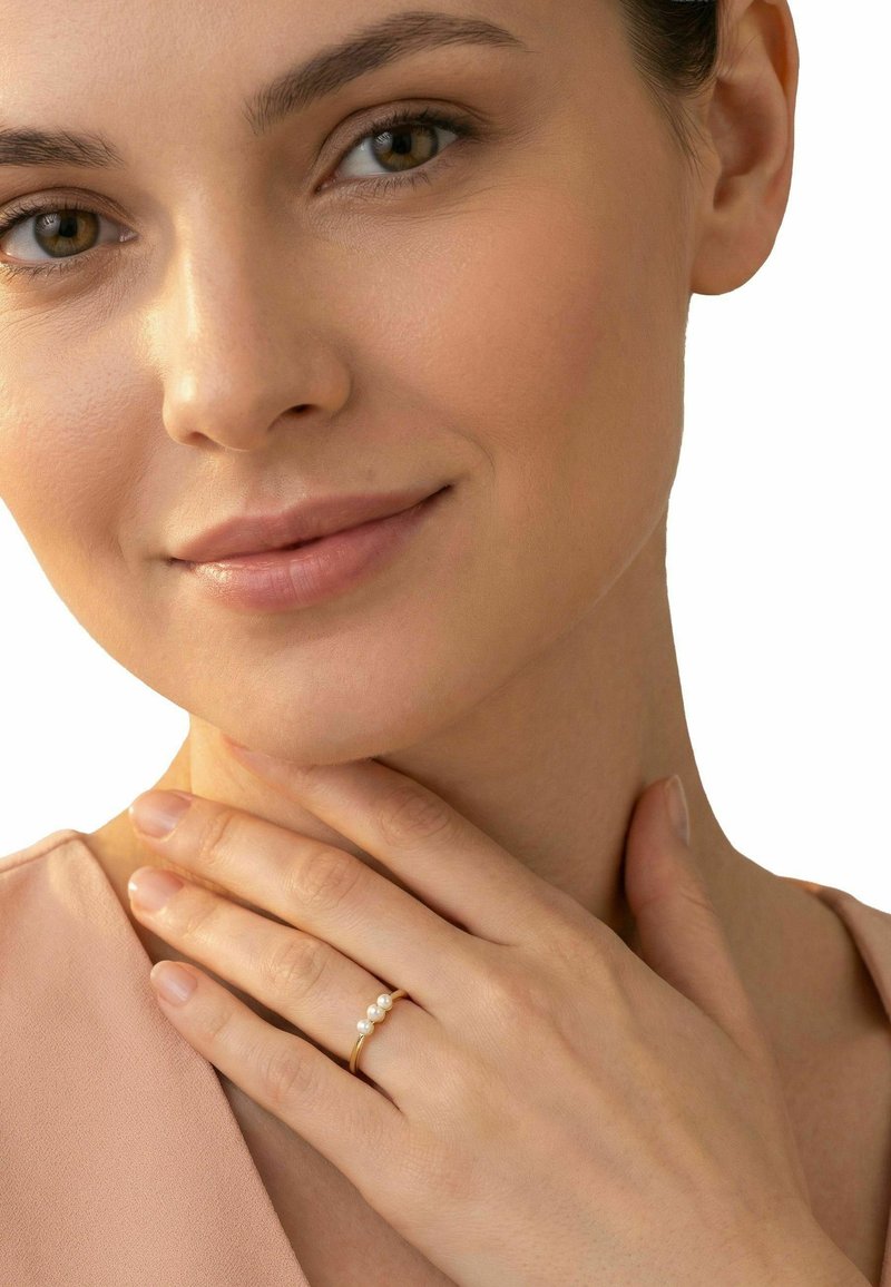Vrouw met een egale huid die zacht haar nek aanraakt, draagt een gouden ring met drie parels aan haar vinger en een lichtroze top.