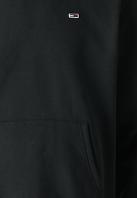 Czarna bluza z kapturem wykonana z mieszanki bawełny, z kieszenią kangurką z przodu oraz małym logo w czerwono-białych i niebieskich akcentach na piersi.