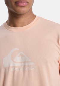 Světle broskvová bavlněná tričko s velkým bílým logem Quiksilver. Má kulatý výstřih a krátké rukávy s hladkou, jednobarevnou texturou.