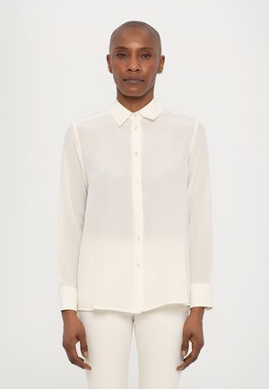 Camicia bianca a maniche lunghe in un tessuto leggero e trasparente, con chiusura a bottoni e colletto classico. Texture liscia e vestibilità su misura.