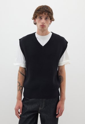 Black sleeveless sweater with a V-neck design, ribbed texture at the edges, layered over a white shirt. Casual fit, worn with dark jeans.