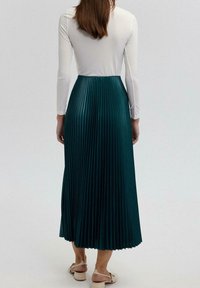 Lange, geplooide rok in donker teal, gecombineerd met een aansluitend wit langemouwen T-shirt. De rok heeft een gladde, gestructureerde afwerking en is enkellang.