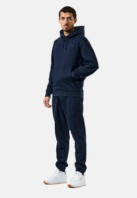 Marineblaues Sweatshirt mit Kapuze, Vorderseite mit Tasche und gerippten Ärmeln; kombiniert mit passender Jogginghose. Vervollständigt mit weißen Sneakern und einer Gummisohle.