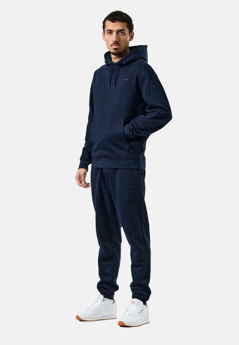 Marineblaues Sweatshirt mit Kapuze, Vorderseite mit Tasche und gerippten Ärmeln; kombiniert mit passender Jogginghose. Vervollständigt mit weißen Sneakern und einer Gummisohle.