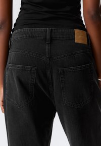 Black denim jeans featuring a high waist, five pockets, and a tan leather patch on the back waistband. Texture is smooth with visible stitching.