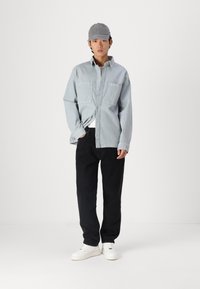Lys blå denimskjorte med to brystlommer, svarte bukser og hvite joggesko. Modellen har på seg en grå caps og står mot en hvit bakgrunn.