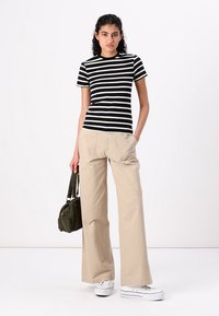 T-shirt a righe bianche e nere con maniche corte abbinato a pantaloni beige a gamba larga. Il modello tiene in mano una borsa verde oliva, indossando sneakers bianche.