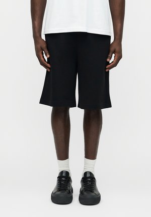 Moški, oblečen v črne kratke hlače do kolen, bele nogavice, črne superge in belo majico, stoji ob navadni ozadju.