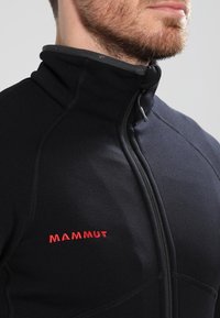 Närbild av en man som bär en svart zip-up fleecejacka med röd "Mammut"-logotyp på bröstet och hög krage.