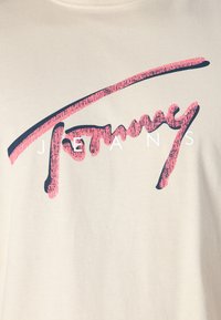 T-shirt crème avec un grand logo stylisé rose portant le mot "Tommy" au-dessus de lettres blanches qui disent "JEANS". Matière en coton avec une texture lisse.
