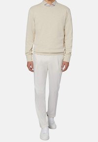 Mann mit beigem Pullover über einem hellgrauen Hemd mit Kragen, weißen Faltenhosen und weißen Sneakers, der vor einem schlichten Hintergrund steht.