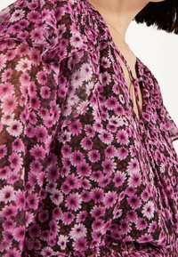 Zbliżenie na przezroczystą różową bluzkę w kwiaty z falbaniastymi rękawami i dekoltem w kształcie litery V, związanym cienkimi sznurkami.