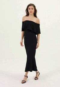 Juodasis off-shoulder suknelė su sluoksniuotu volanu, prigludusi siluetas ir kulkšnies ilgio sijonas. Dėvima su juodomis juostelėmis sandalais.