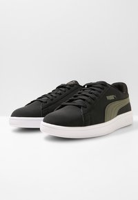Zapatillas sintéticas negras con un acabado suave, que presentan una franja de acento verde oscuro, suela plana de goma blanca y sistema de cordones estándar.