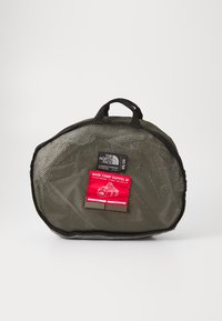 Mesh duffel bag in dark green with black accents. Features a red label detailing capacity and dimensions. Rounded shape, dual top handles.