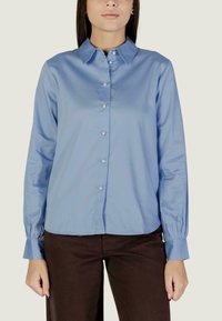 Donna che indossa una camicia azzurro chiaro con bottoni, colletto e maniche lunghe, abbinata a pantaloni marrone scuro, in piedi davanti a uno sfondo neutro.